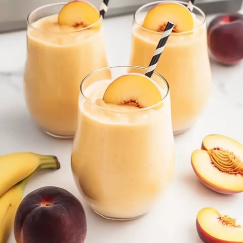 Three glasses of creamy peach and banana drinks topped with fresh peach slices, striped straws, and surrounded by fruit.