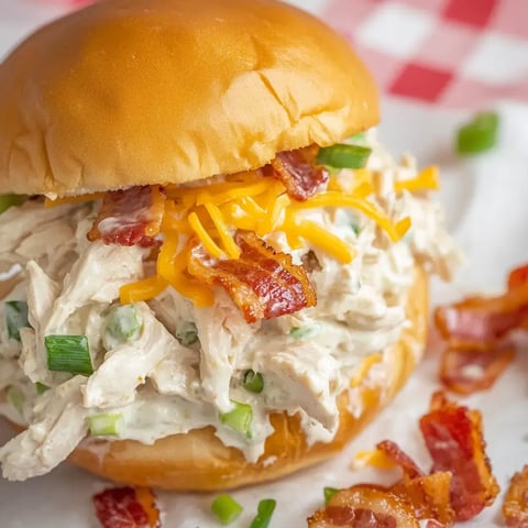 Close-up of shredded chicken with bacon and cheese on a bun, topped with green onions and served with crispy bacon bits.