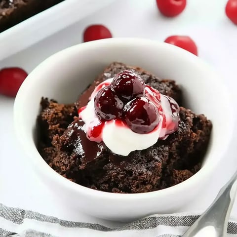 A dish filled with layers of chocolatey cake, cherries, whipped cream, and a drizzle of cherry sauce, with fresh cherries scattered around.