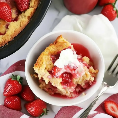 A slice of warm strawberry cobbler with whipped cream, beside fresh berries on a striped cloth.