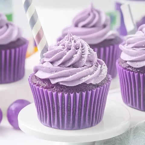 Close-up of pretty purple cupcakes with swirled icing and striped straws, resting on a white stand.