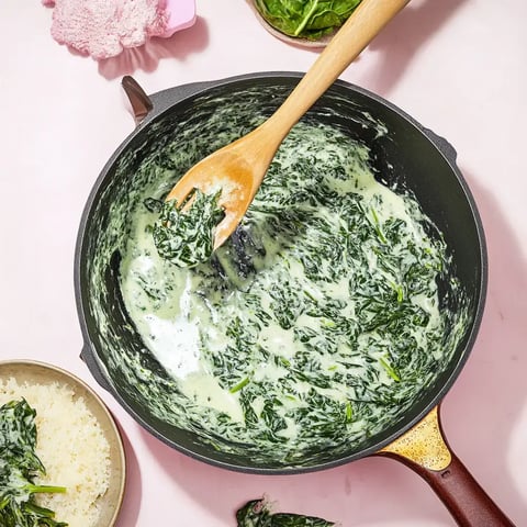 A black pot with creamy spinach being stirred by a wooden spoon, surrounded by rice and spinach leaves on a pink surface.