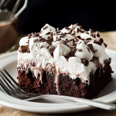 A rich chocolate cake topped with fluffy whipped cream, curls of chocolate, and soft marshmallows on a white plate with a fork.