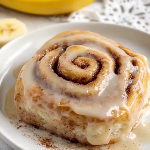 A stack of warm banana cinnamon rolls covered in frosting with a cinnamon sprinkle on top.